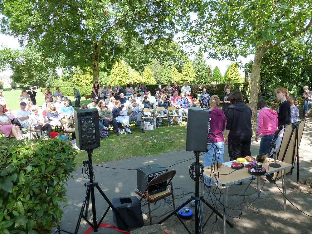 This photograph is taken from just in front of the murals facing the audience. It is a very sunny day.  There is a crowd sitting under the shade of trees in the park. A group of 5 participants is standing at the front performing to the audience. There is equipment for amplifying the sound to the left of the performers. 