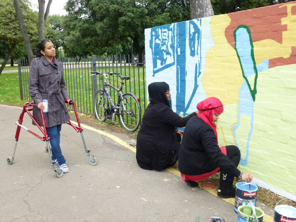 There are two women crouching down painting the mural, they are both wearing headscarves. They are painting a figure in yellow and a figure in light blue. A woman walking with a frame is watching them. She is wearing jeans and a checked jacket and has dark hair tied back in a pony-tail.