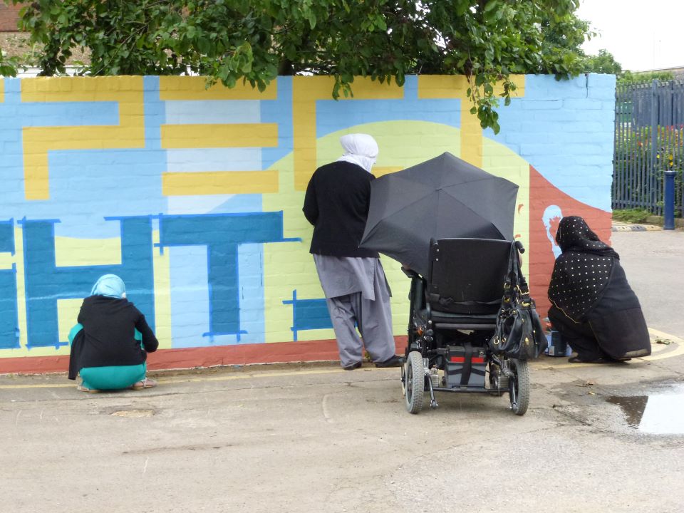 Four women are painting on the wall. They are painting the background and the letter PECT in yellow and HTS in blue from the words RESPECT and RIGHTS. Two women are crouching down, they are wearing headscarves, one woman is a wheelchair user and is holding an umbrella and the other woman is standing up to paint.