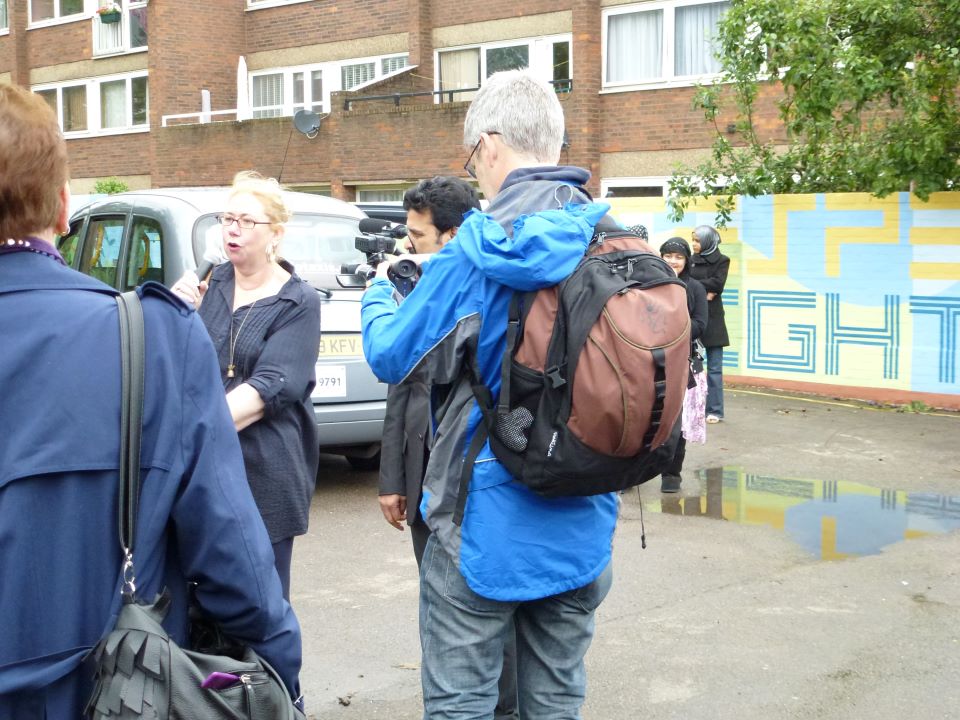 This is a photo of the opening event. A lady who has long blonde hair in a pony-tail and is wearing glasses is speaking to the crowd and someone is filming her. In the background you can see the mural.