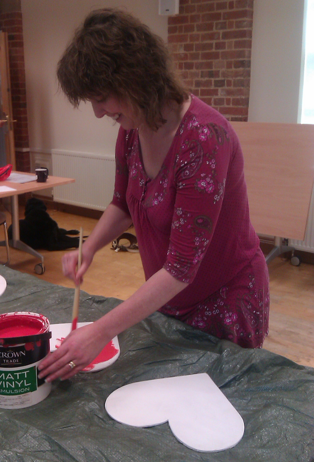 A woman is  smiling as she paints red paint on a white small piece of wood. The shape of the wood is hidden behind the paint pot. The woman has long wavy hair. She is wearing a pink top with flowers on it.
