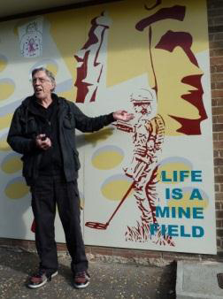 This picture shows a man wearing black jacket and trousers. He is standing with one arm pointing towards the line drawing of the soldier mine sweeping.