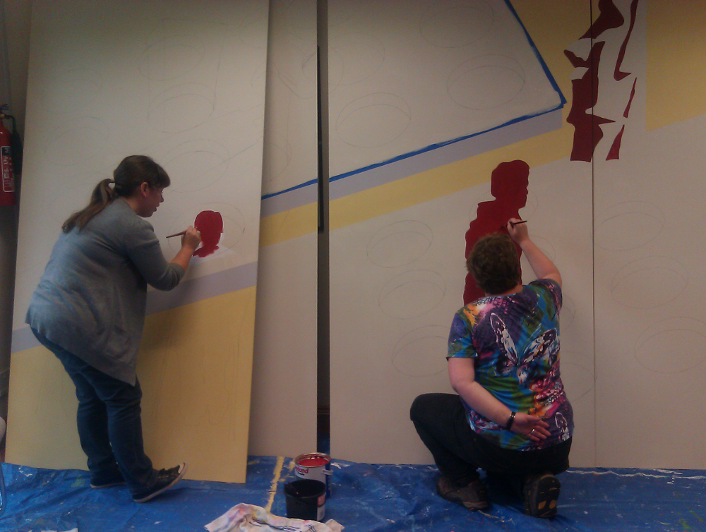 This photo shows two women painting on boards that are propped against the wall. On the left there is  a woman with long dark hair tied in a single bunch. She is wearing a grey cardigan and blue jeans. Next to here there is another woman with one hand behind her back. She is wearing a brightly coloured t shirt and black trousers. She is kneeling on a blue tarpaulin on the ground. They are both painting with dark red shadows of figures against a background with cream, yellow and grey.