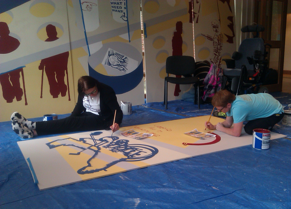 This pictures shows two people painting on a board on the floor. One woman is sitting on the floor with her legs straight in front of her.  She is dressed in black trousers, black cardigan, white t shirt with a drawing on it, and black and white slippers. She is painting the dark blue outline of someone in a wheelchair. The other woman is kneeling and bent over the board as she is painting. She is wearing a light blue t shirt and black trousers. She is painting a dark red semi-circle. Both women are wearing glasses. At the back there are two more boards propped against the wall.  These are the boards for the long wall of the mural in Norwich. There are dark red figures using crutches with their heads sticking out of circular holes, next to them a bit lower down is a dark blue circular bin with a picture of a screaming face inside it. On the right there is a pile of chairs hiding most of the painted boards.