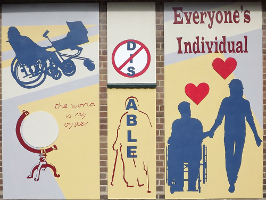 The mural is in three panels. On the right of the mural are the words \'Everyone\'s individual\'. Below this are dark blue silhouettes of a wheelchair user holding hands with someone standing up. Red hearts are above their heads. The middle panel has the letters \'DIS\' crossed out and beneath it the word \'ABLE\' written vertically down the outline of a person using crutches. On the left there is a blue silhouette of a wheelchair user pushing a pram. Underneath this is a picture of a globe with the words \'The world is my oyster\'.'